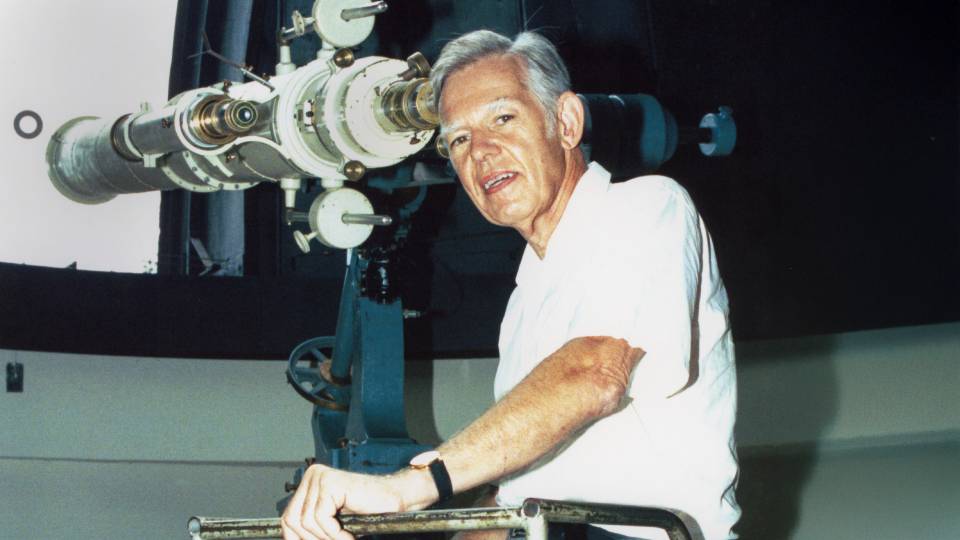 A white-haired man glances down from a massive telescope.