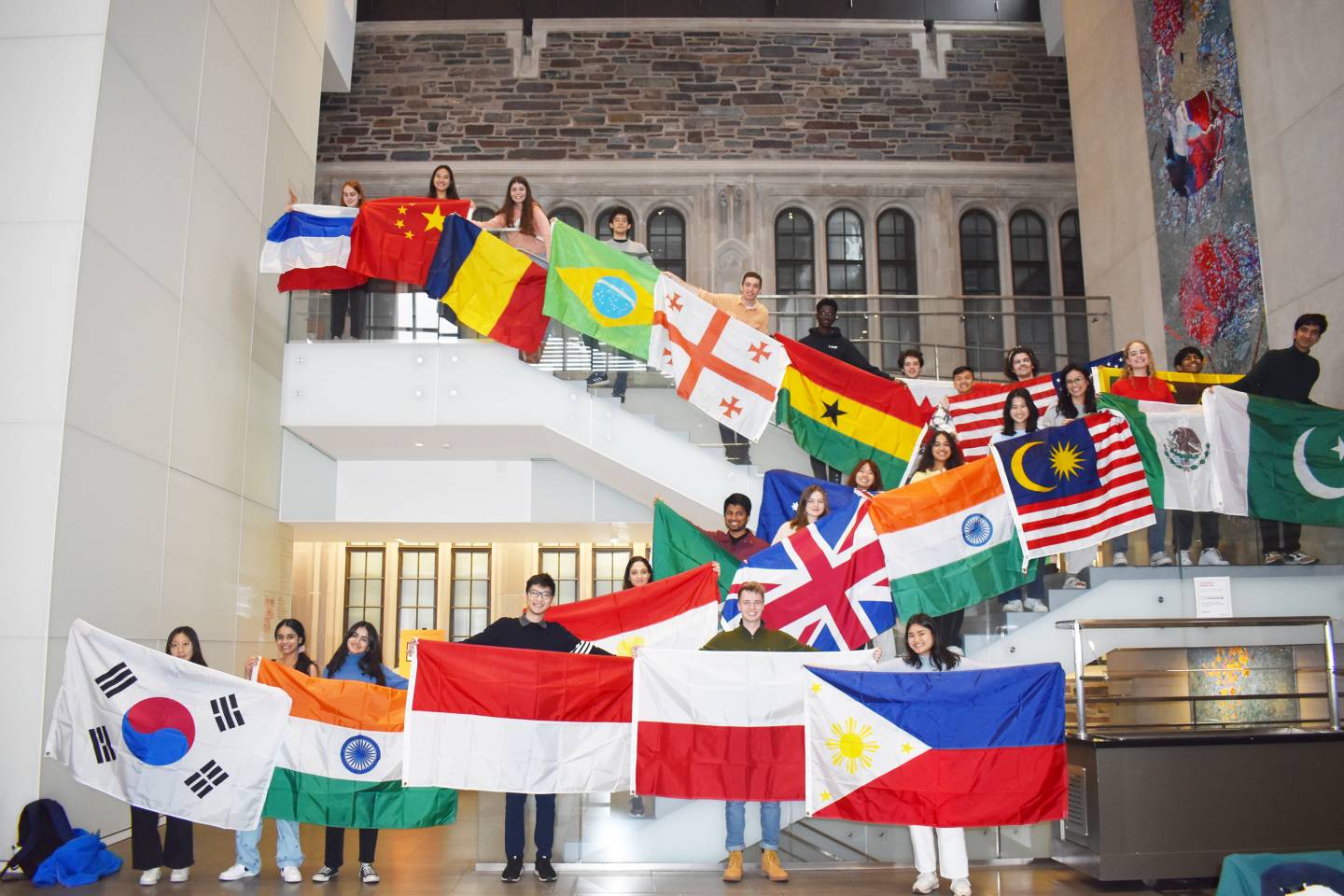 International Students holding the flags of their home nations pose on a stairway inside the Simpson International Building