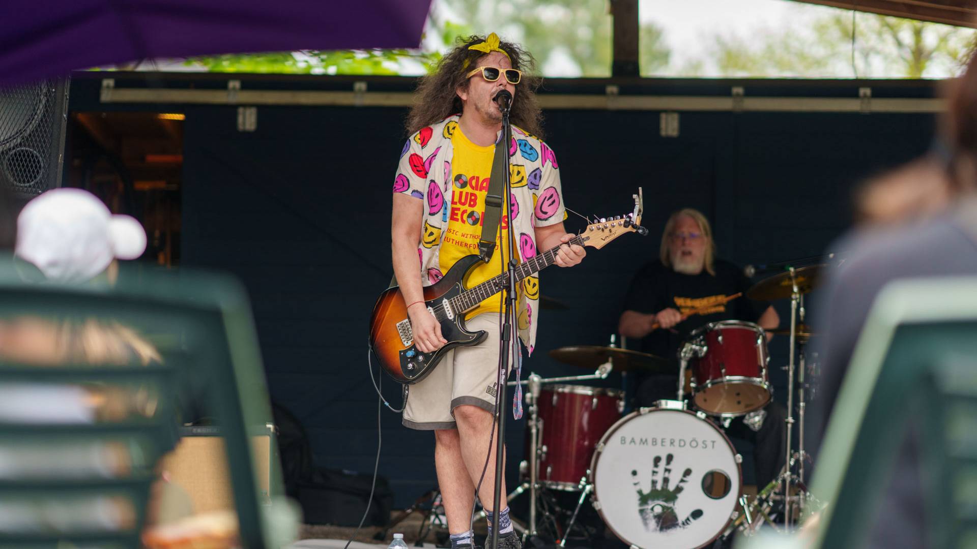 A guitarist in a bright yellow-pink ensemble sings outside a local home with a drummer in the background.