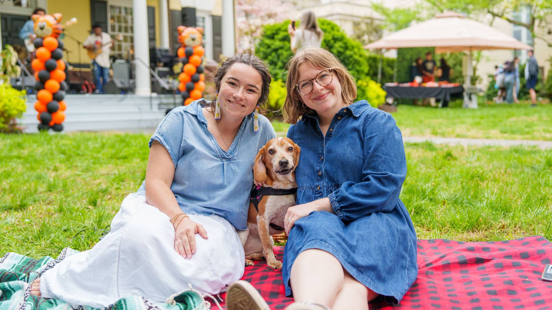 Two students sit on a blanket on the lawn outside Maclean House with their dog in the middle.