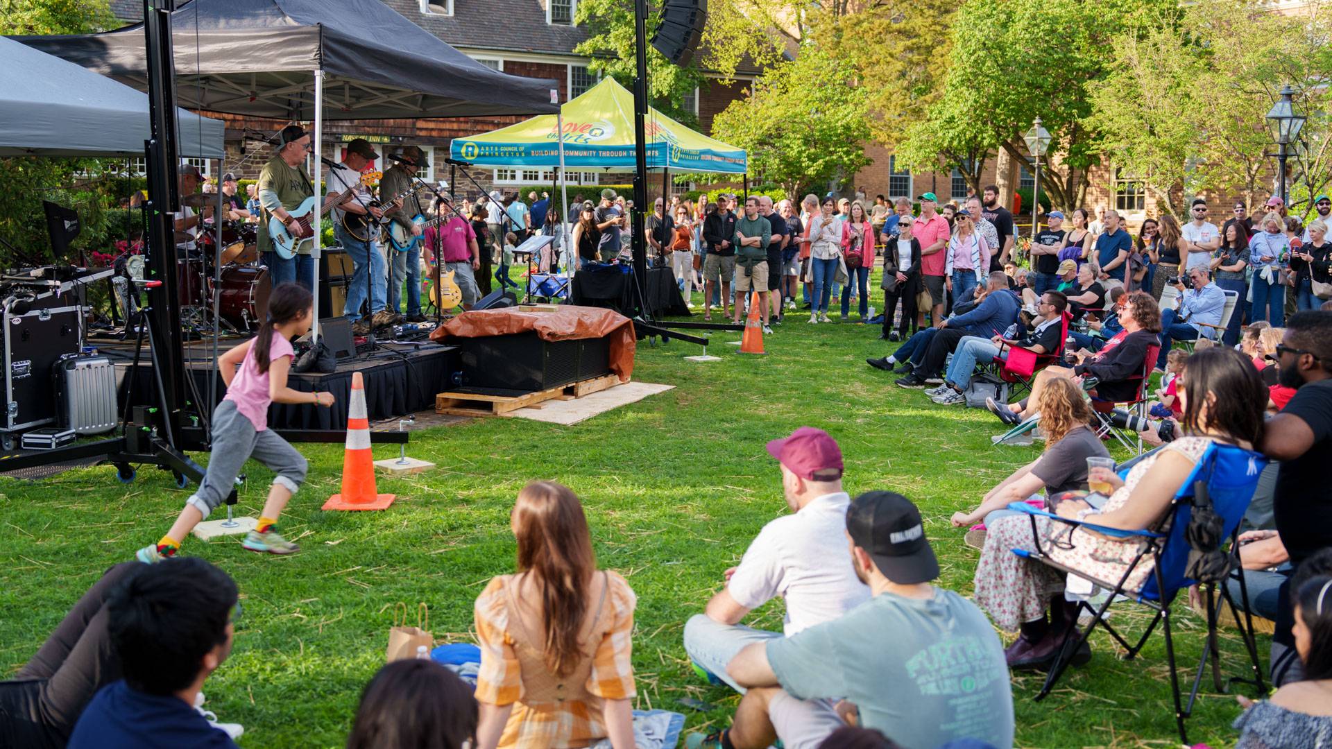 An attentive crowd sits around an outdoor stage to enjoy a bright summer evening of music.