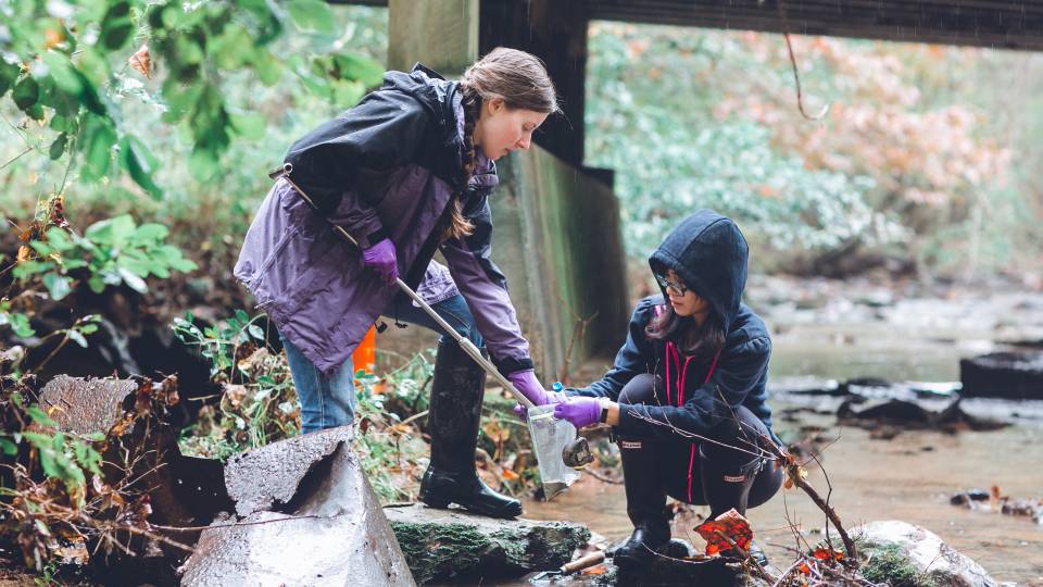 Two students collecting water samples from the bank of a river