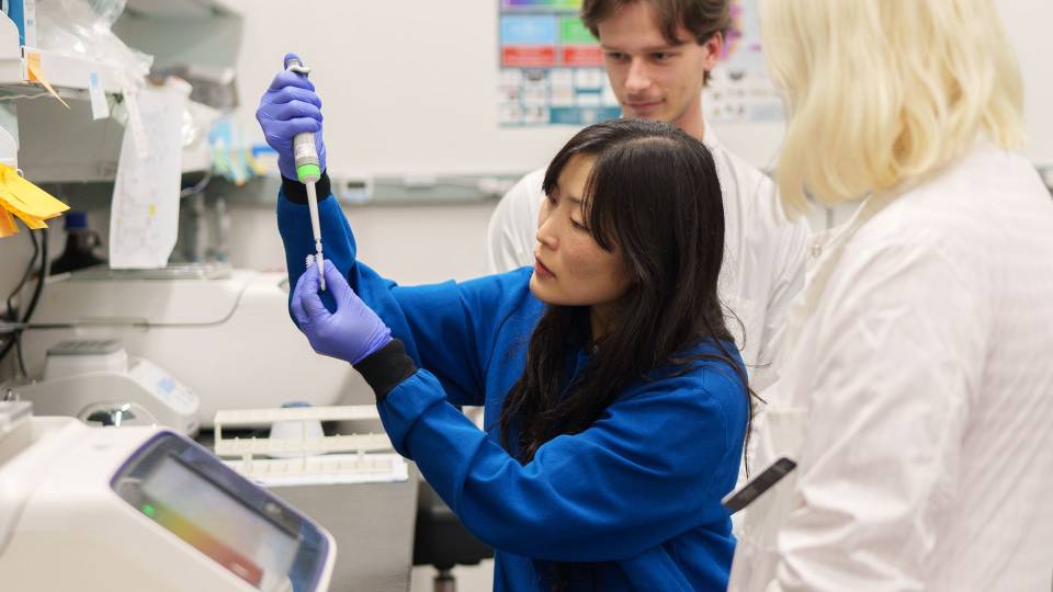 A woman pipettes a fluid into a tiny container while students look on.