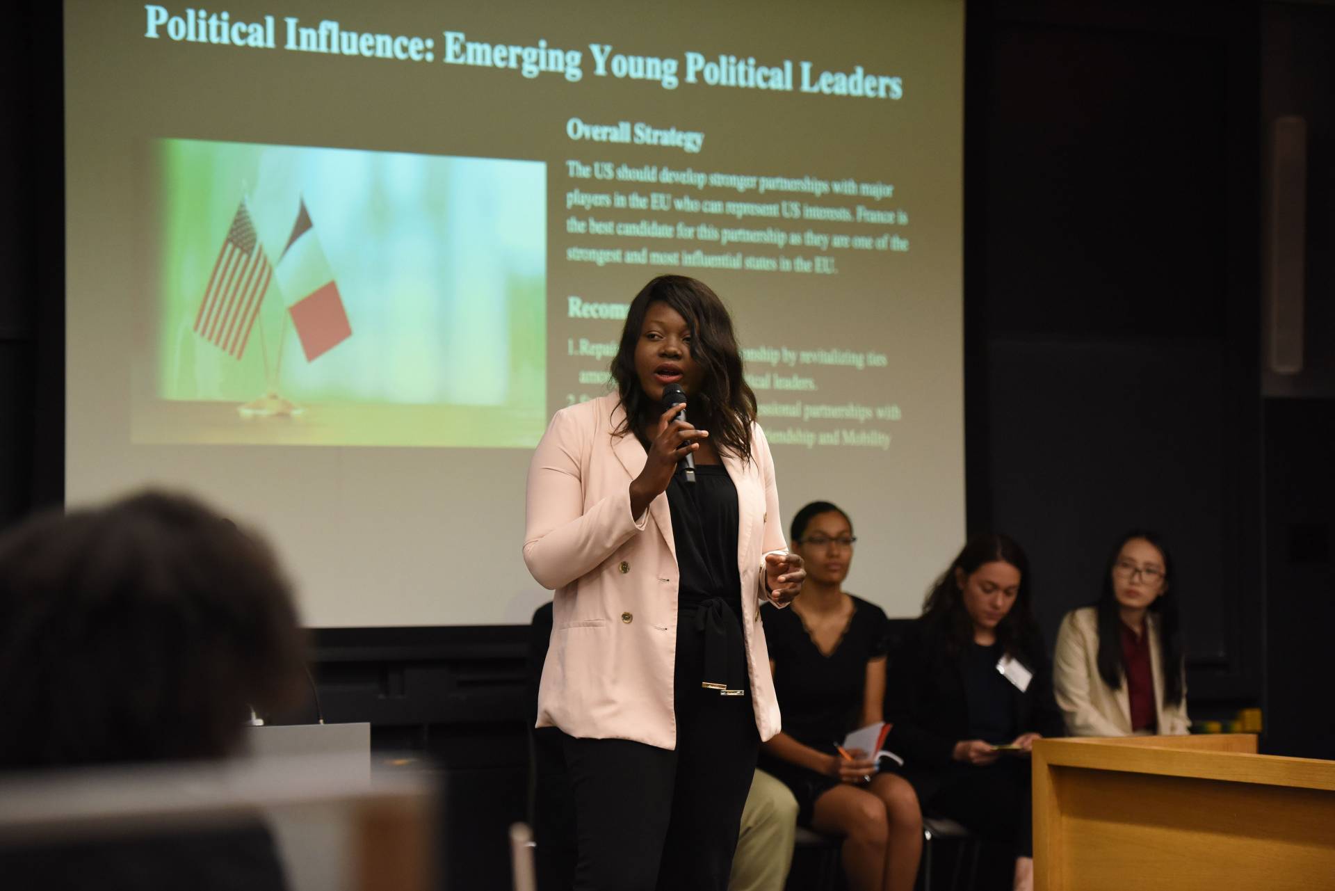 A woman speaks to an auditorium-style classroom full of undergraduates in front of a projection that reads: "Political Influence: Emerging Young Political Leaders" 