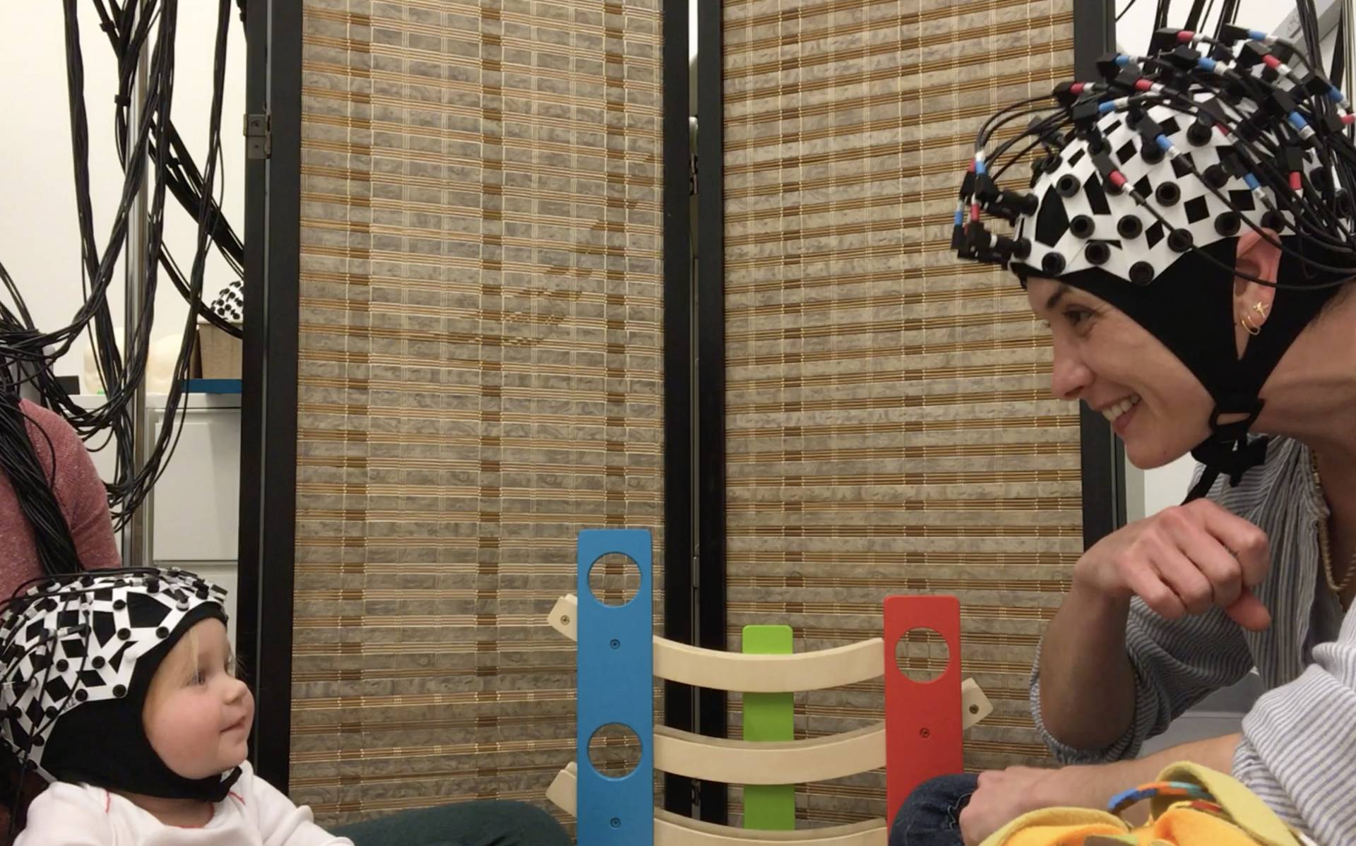 A baby and a researcher wear brain wavelength-measuring devises on their heads while making eyecontact during play