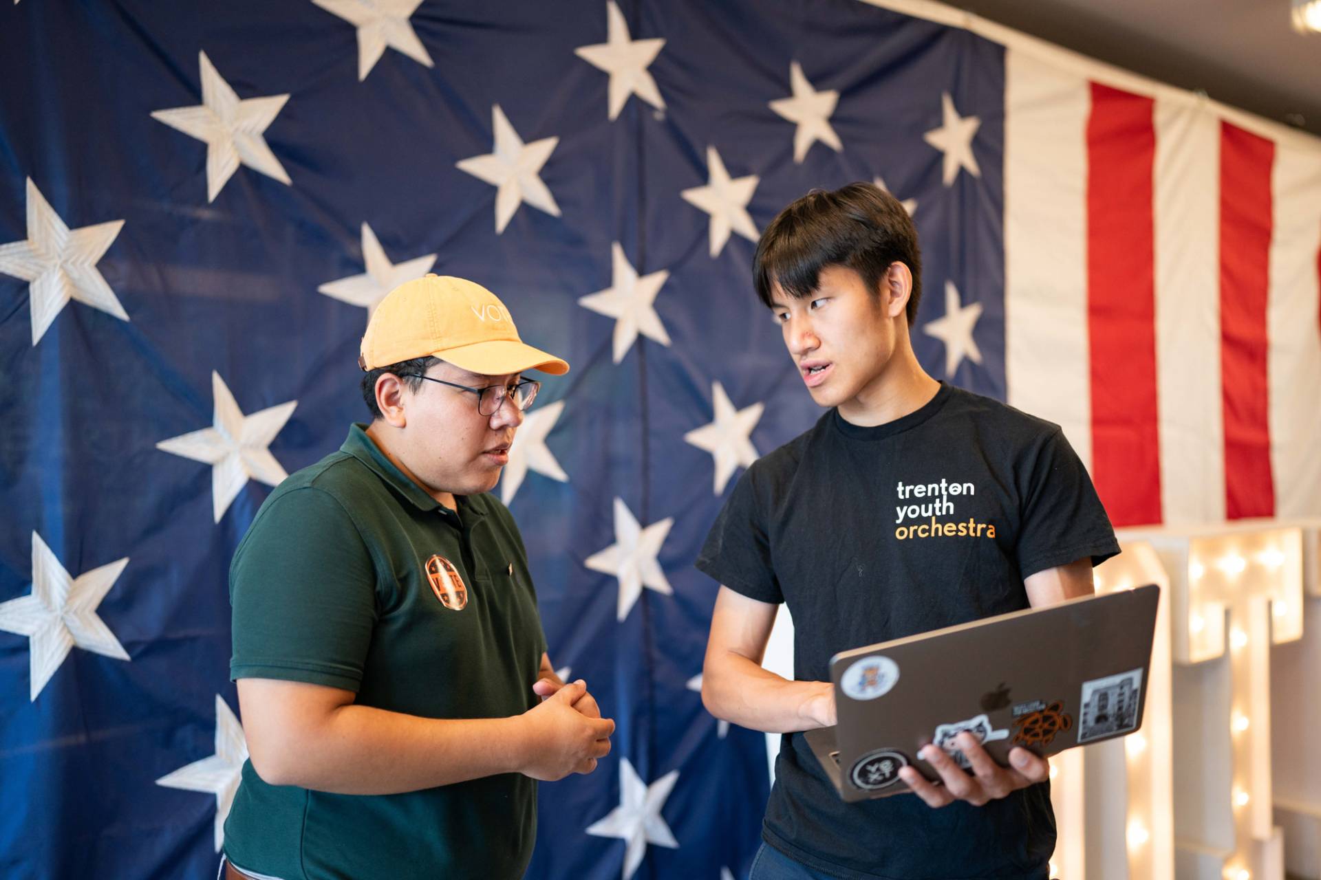 A person holding a laptop talking to another person with the US flag behind them.