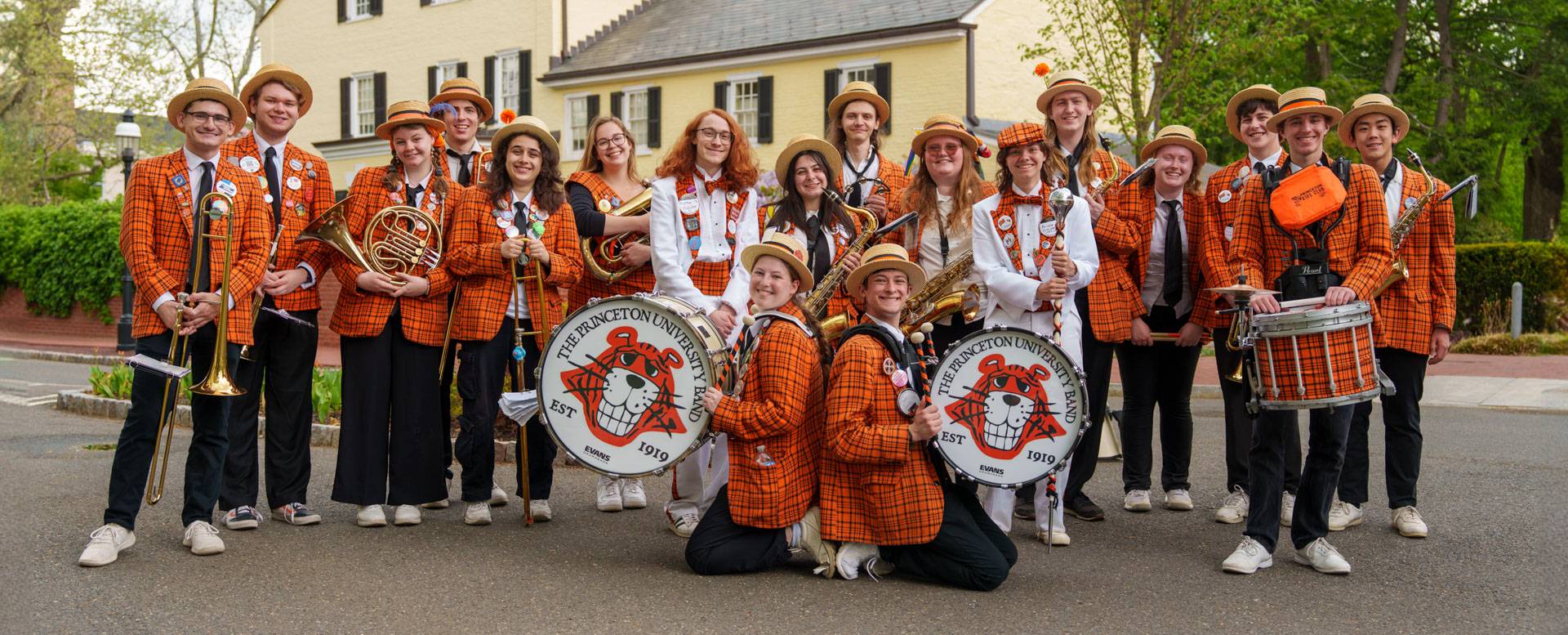 A large student band, most clad in orange plaid suit jackets, stops for a group photo holding their instruments.