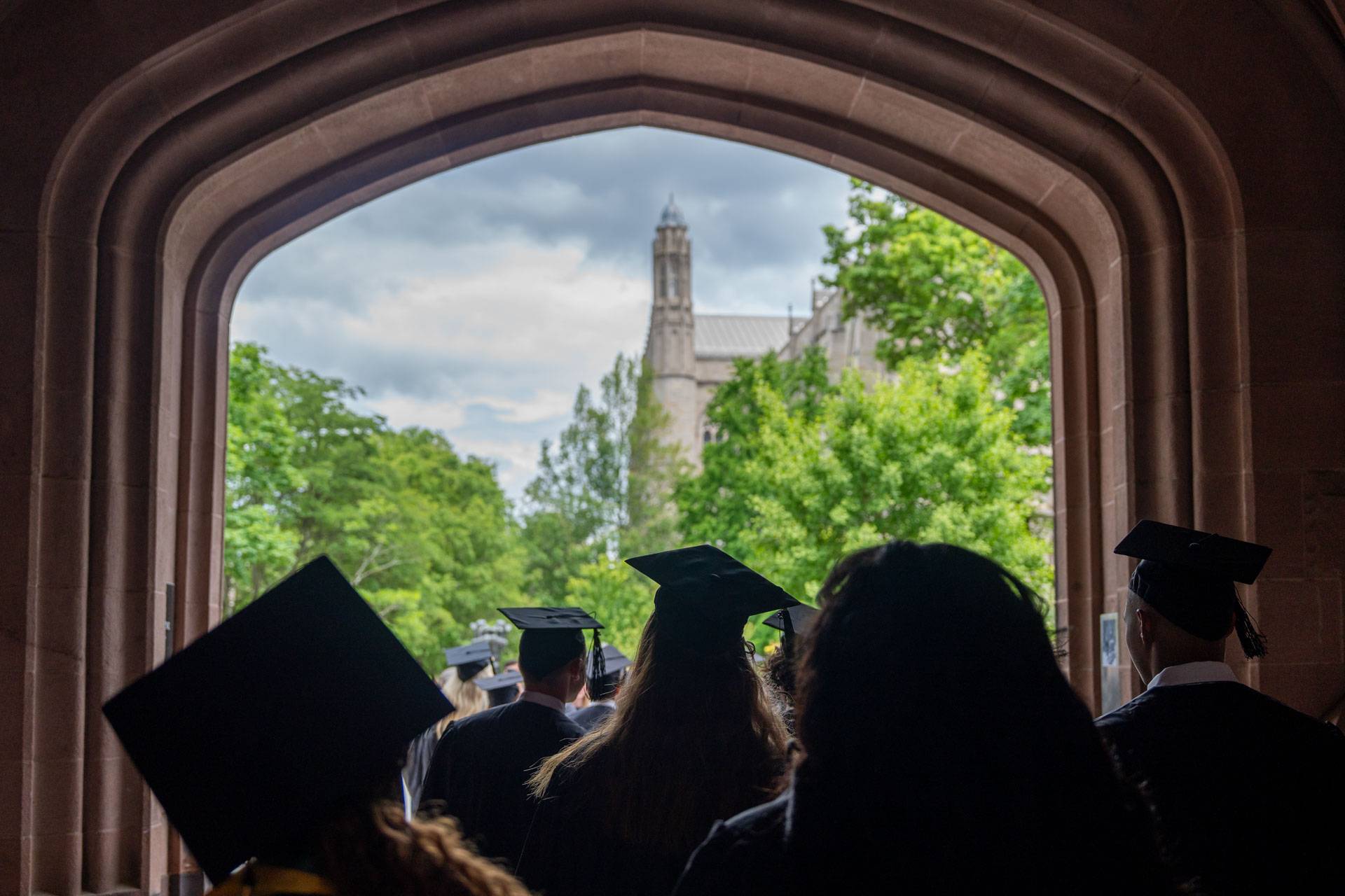 Graduating seniors in their caps and gowns walk through an archway.